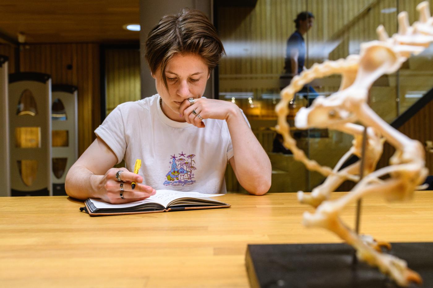Een persoon zit met een pen in de hand en een opengeslagen schrift aan tafel. Rechts in beeld staat een deel van een skelet van een dier. Op de achtergrond loopt iemand de trap op.