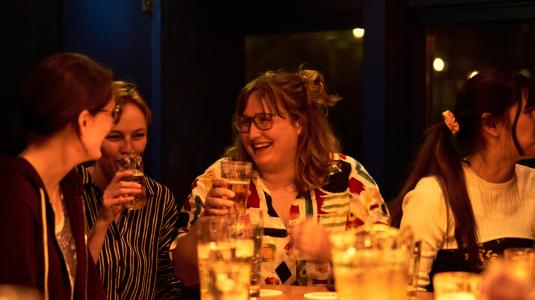 Vier studenten zitten aan de bar met drankjes voor hun op tafel en biertjes in hun hand. Ze lachen en hebben plezier.